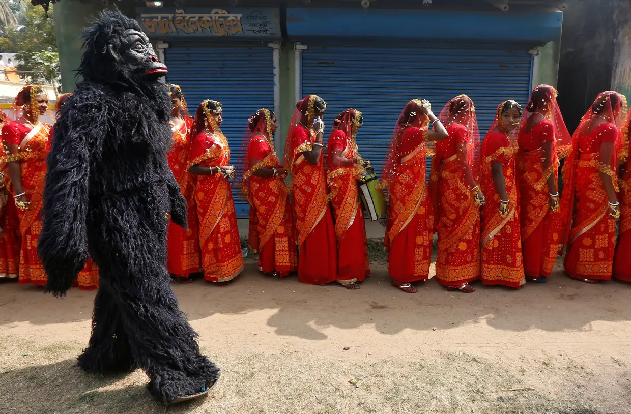 Figurante vestido de chimpanzé passa junto a uma fila de noivas que se preparam para a cerimónia que realiza em simultâneo o casamento de mais de cem casais muçulmanos e hindus próximo de Calcutá, Índia.