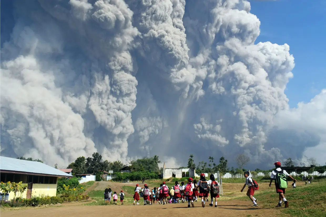 Os estudantes indonésios caminham juntos na escola primária de Sipandak, na aldeia de Tiga Pancur, em Karo, Sumatra do Norte, em 19 de fevereiro de 2018, quando cinzas vulcânicas espessas do vulcão Monte Sinabung se elevam no ar depois de outra erupção