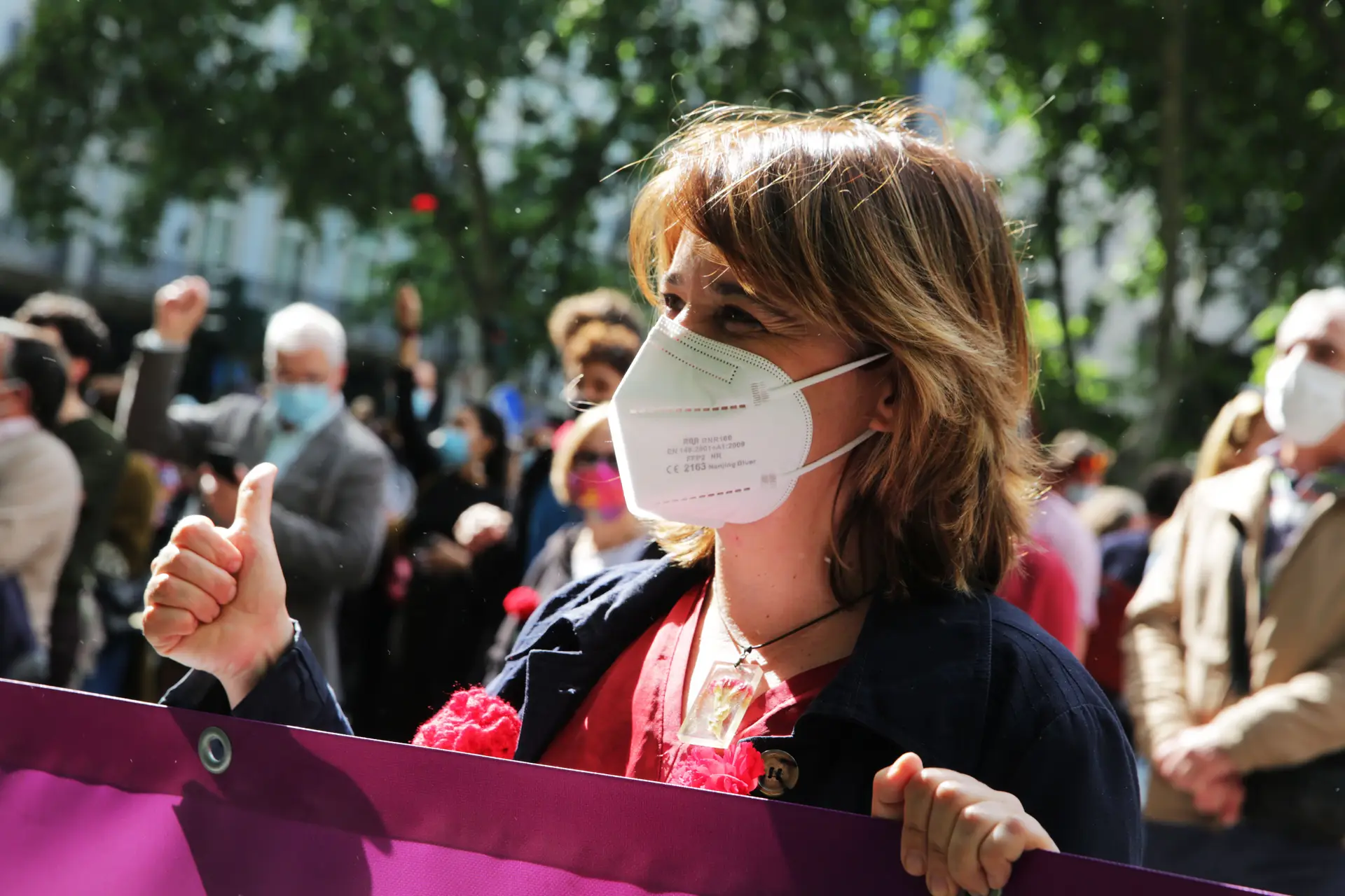 Catarina Martins com uma camisa vermelha e um cravo no bolso do peito do casaco. A máscara branca esconde o rosto da líder bloquista, mas os olhos exibem o sorriso.