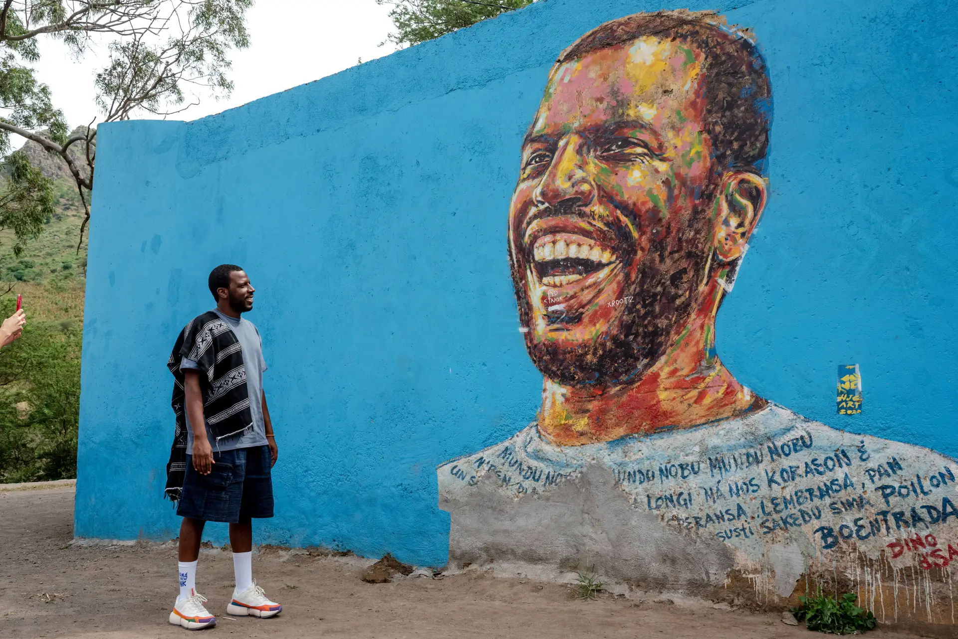 Dino D'Santiago junto a um retrato seu feito na parede da escola, junto a Boentrada, a terra do pai.