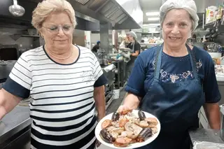 Rosa Quesado com olhar sempre atento aos pratos que saem da cozinha. Judite Quesado segura uma travessa com carnes da salgadeira e enchidos
