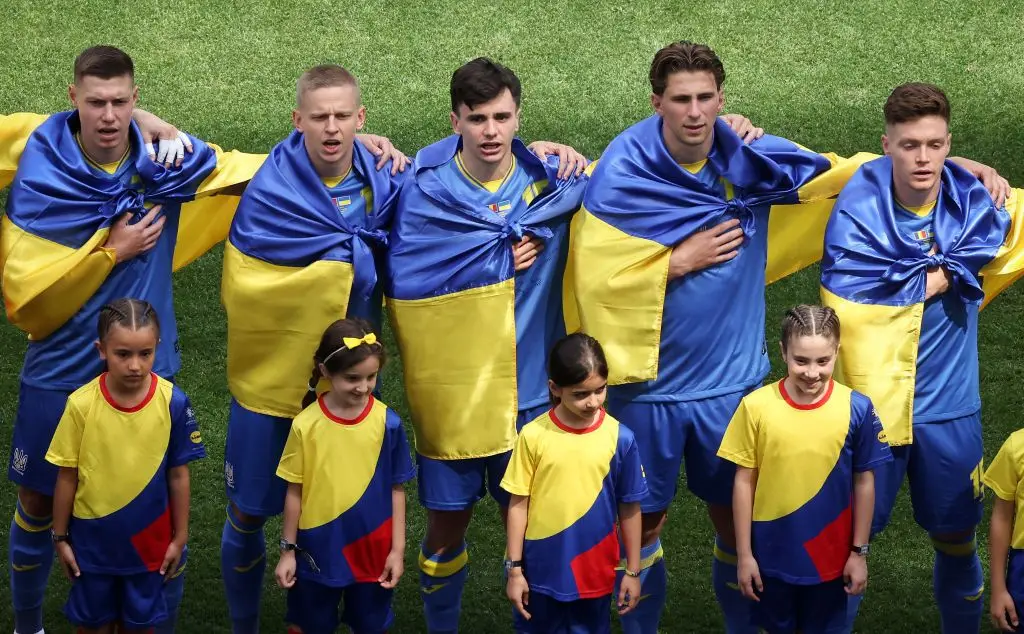 Jogadores da Ucrânia escutam o hino, antes do jogo com a Roménia, embrulhados em bandeiras do país, em guerra há mais de dois anos