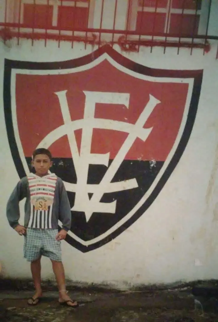 O médio brasileiro aos 11 anos, junto do emblema do Vitória da Bahia, primeiro clube onde jogou