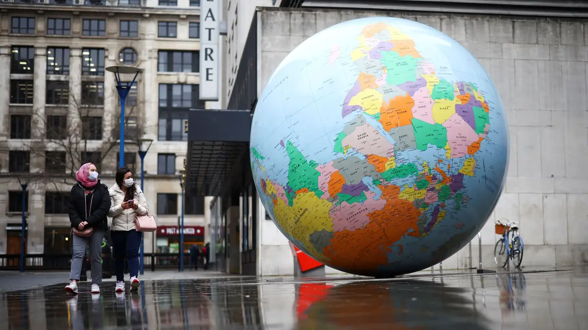 Pessoas com máscara de proteção junto à escultura "The World Turned Upside Down", de Mark Wallinger, em Londres, Inglaterra.