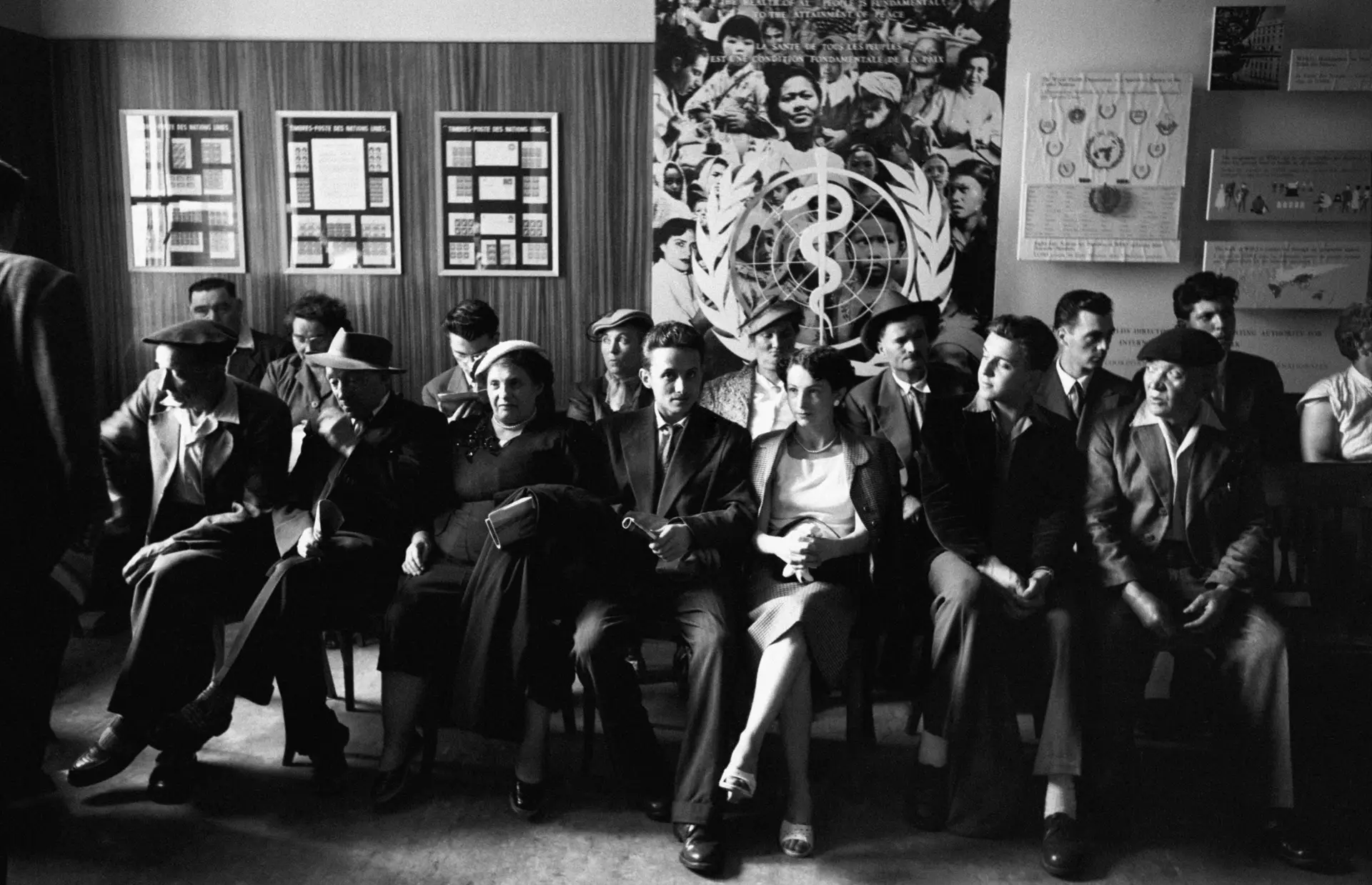 Pessoas sentadas em frente a um cartaz da Organização Mundial da Saúde em Genebra, cidade suíça que alberga a sede da agência da ONU fundada em 1948. A foto é de uma reunião, realizada em 1955, para debater questões de segurança, reunificação alemã e desarmamento.