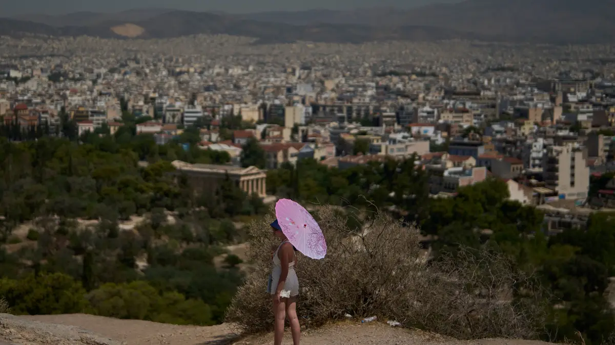 Uma turista com um guarda-chuva está em pé na colina Filopapo, com Atenas estendendo-se ao fundo, segunda-feira, 7 de julho de 2025.